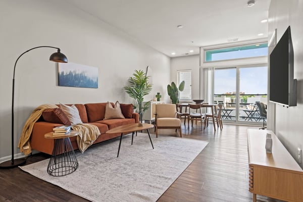 Modern living room with rust-colored sofa, arc lamp, potted plants, and floor-to-ceiling windows overlooking a balcony with city views.